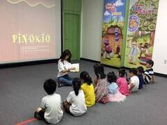9명의 어린이들이 나란히 바닥에 앉아서 책을 손에 들고 설명하고 있는 강사의 말에 경청하고 있는 모습