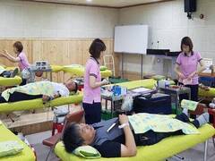 건물내 임시로 간이 침대가 여러개 놓여져 있으며 침대 위에 헌혈을 하고 있는 남성들이 있으며 분홍색 유니폼을 헌혈 담당자들이 헌혈 업무를 하고 있는 모습 
