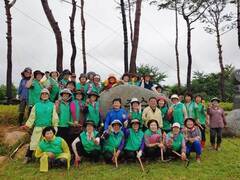 관산읍 지역 새마을남녀지도자 40여명이 초록색 조끼와 모자를 쓰고  초록 잔디밭 위 기념석이 있는 곳에서 한손을 들고 화이팅 자세로 단체 기념 촬영을 하고 있는 모습