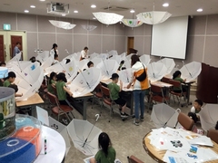 물과학관 교실에 초록색 반팔티와 검은 바지를 입은 단체 어린 학생들이 의자에 앉아서 책상위에 놓여진 하얀색 반투명 우산에 그림을 그리고 색칠을 하고 있으며, 강사들이 돌아다니며 아이들을 지도하고 있는 모습