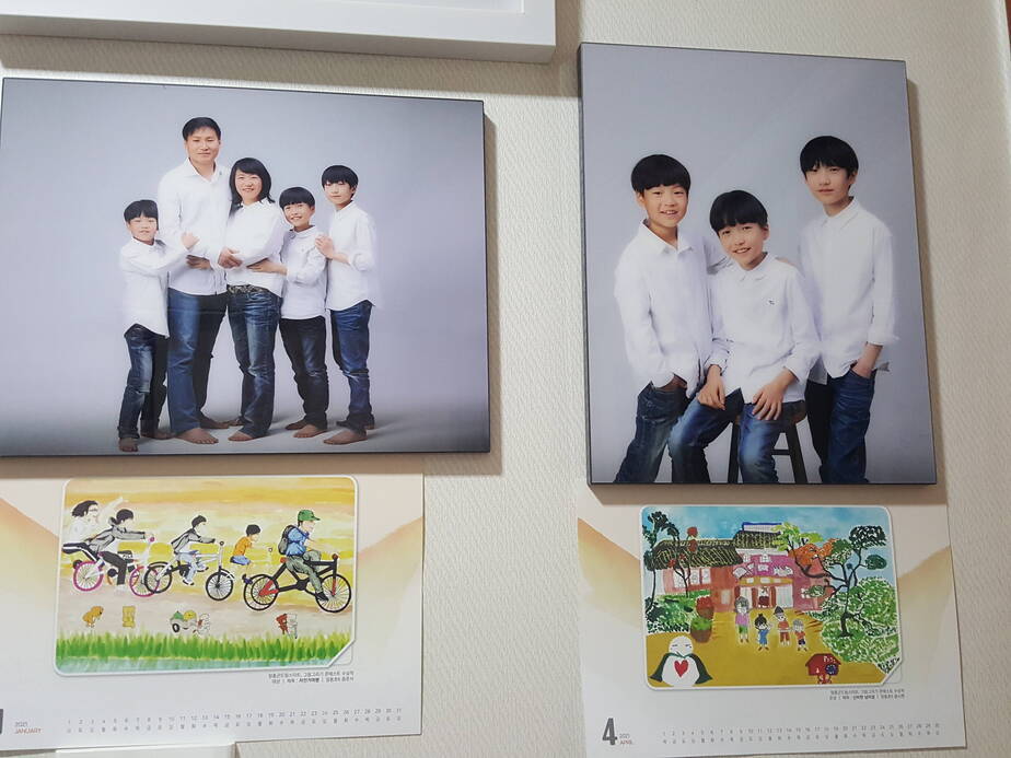 벽에 가족 사진과 그림이 있는 달력이 붙어 있다. 왼쪽 사진에는 엄마 아빠 아들 3명이 흰 티셔츠와 청바지를 맞춰 입고 나란히 서 있고 오른쪽 사진은 같은 옷을 입은 아들 3명이 함께 찍은 모습이다.