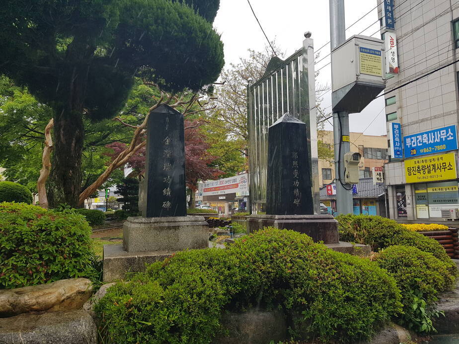 두 비석을 위 쪽 사진보다 조금 더 멀리서 찍은 사진이다. 비석 왼쪽에 커다란 나무가 있고 오른쪽 도로 옆에는 빌딩이 있다.